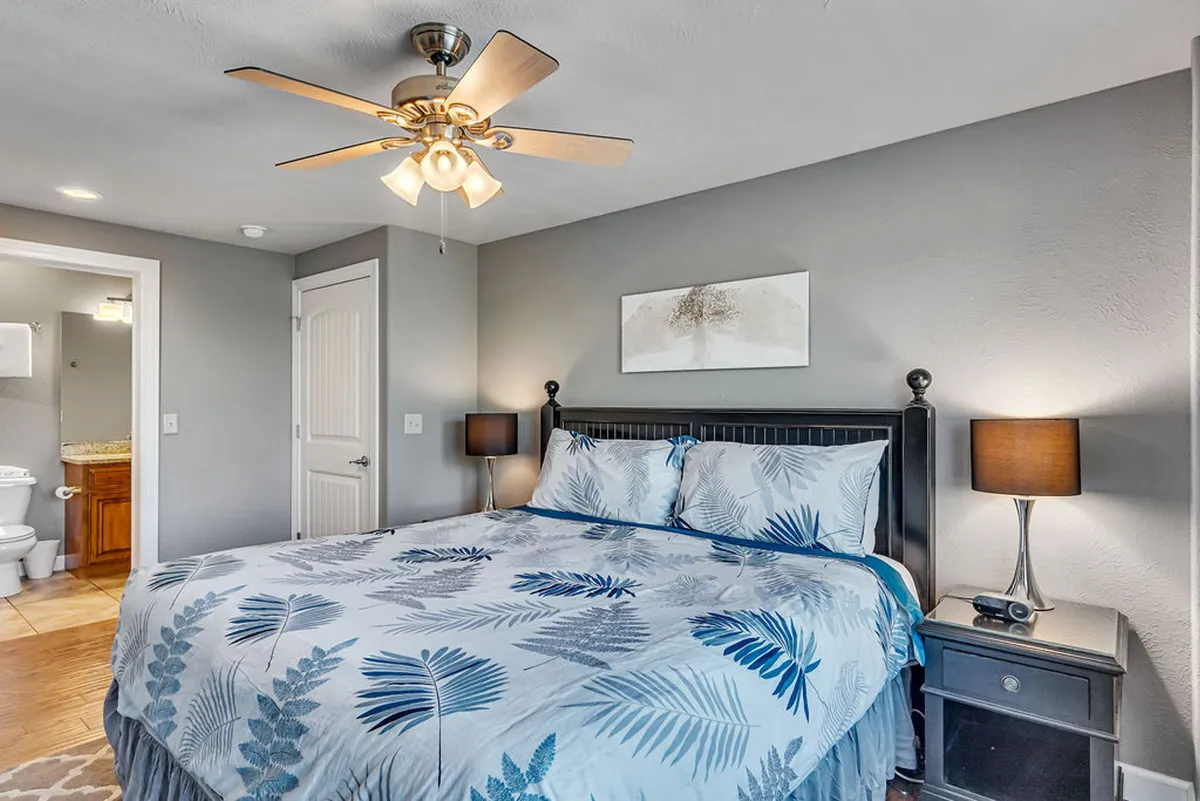 A serene and comfortable king bedroom featuring blue and white leaf-patterned bedding, matching nightstands with lamps on either side of the bed, and a ceiling fan overhead. Natural light filters softly through the windows, creating a restful and inviting atmosphere.