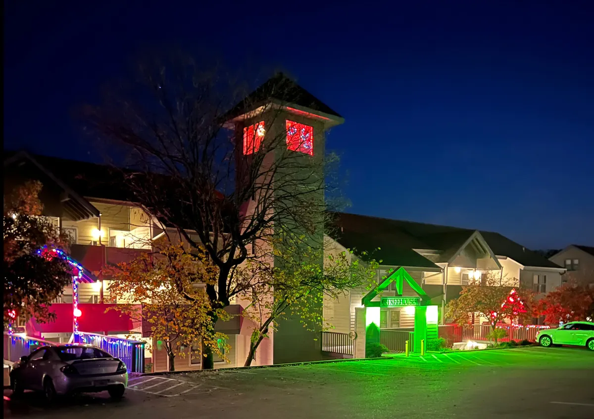 An evening exterior shot showcasing a clock tower, green entrance sign, and trees decorated with glowing lights. The area is bustling with the cheerful ambiance of community gatherings, highlighting both the property’s inviting entrance and its festive neighborhood surroundings.