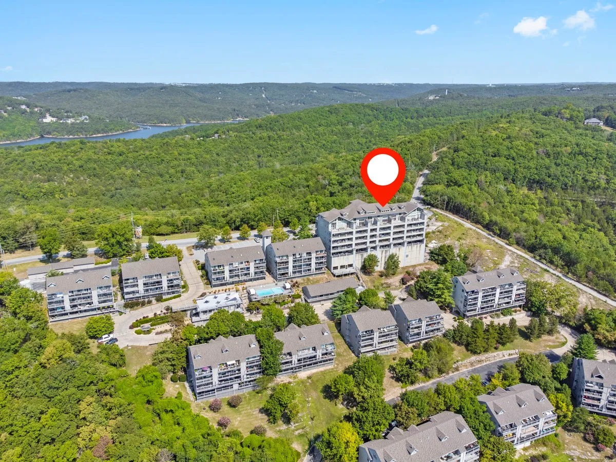 This image displays the exterior of a multi-unit residential complex surrounded by landscaped greenery, with a sparkling swimming pool as the central focal point. Lounge chairs line the pool, and the tranquil setting is complemented by the presence of trees and inviting walkways.