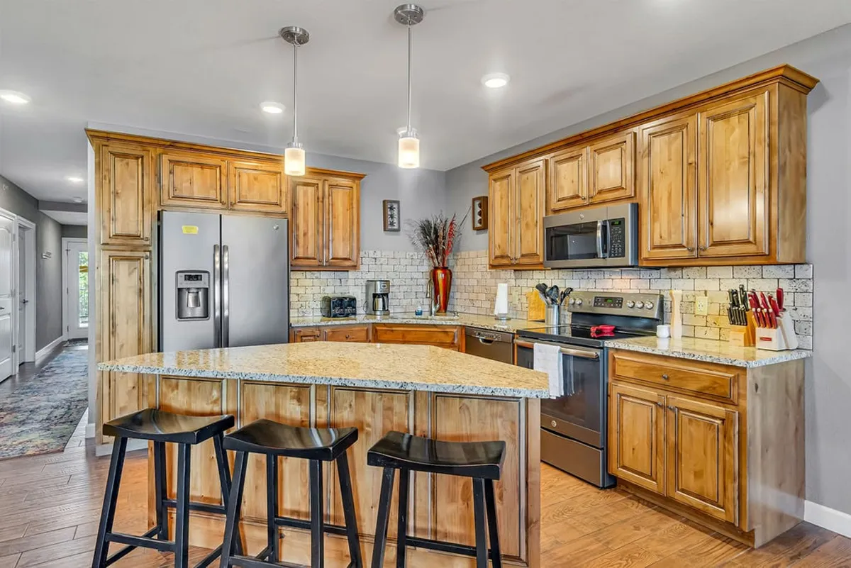 A warm and inviting kitchen scene featuring an expansive granite countertop, island seating, wooden cabinetry, and high-end appliances. The setting hints at lively gatherings with friends or family, perfect for entertaining and socializing.