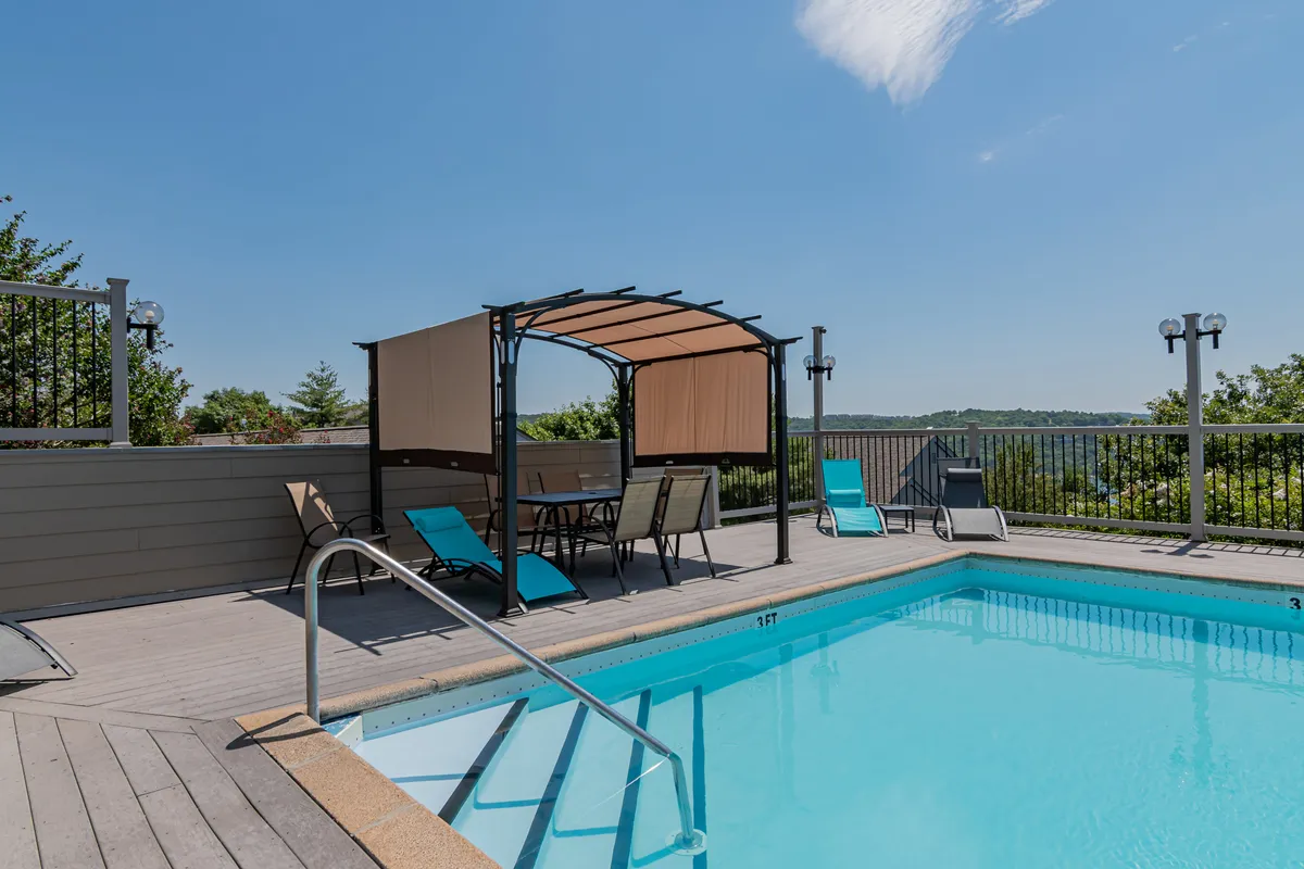 The image features a sunlit swimming pool framed by a canopy seating area and lush landscaping, offering a scenic spot for swimming, sunbathing, and socializing. The tranquil background highlights the resort feel of the community.