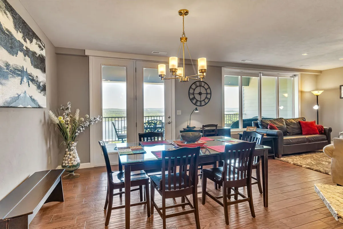 A view of an inviting dining table set beneath a chandelier, with a sofa and large windows in the background. Artwork adds character, and the windows offer glimpses of the scenery outside. The arrangement suggests a space ideal for shared meals and relaxation.