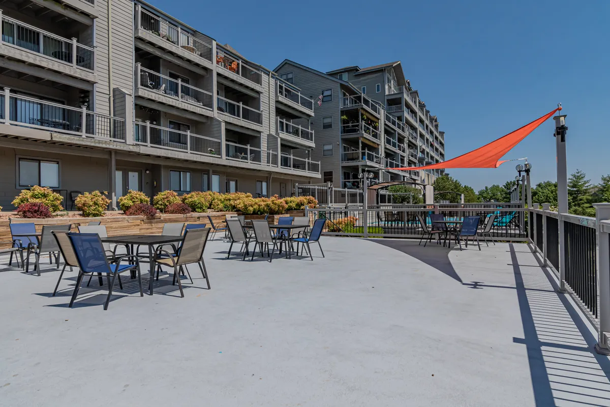 An inviting outdoor seating area with flower beds, a stylish shade sail, and the building's architecture in the background. The space suggests opportunities for socializing, relaxing, or enjoying a morning coffee with a view of the surrounding landscape.
