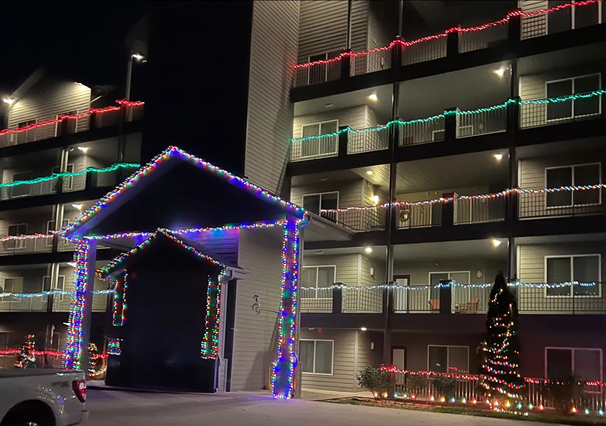 A warmly lit exterior of a condo building adorned with festive holiday lights. The entrance is welcoming, with balconies illuminated and a holiday tree set up near the entryway. The scene conveys a sense of arrival and community spirit, suggesting a lively and inviting environment.